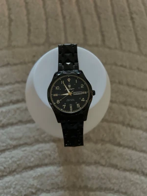 Svart klocka med pyramidmönstrat armband - Inte copy hög kvalitet och ny rekommenderad för still. Stilren svart klocka med rund urtavla och guldfärgade visare. Armbandet har ett unikt pyramidmönster som ger en cool och modern look. Urtavlan visar både dag och datum. Perfekt accessoar för dig som vill sticka ut.