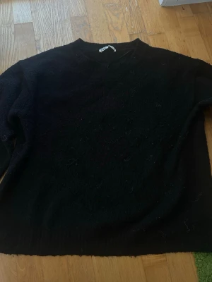 Svart stickad tröja med rund hals - En enkel och stilren svart stickad tröja med rund hals och långa ärmar. Tröjan har ribbstickade muddar vid ärmslut och nederkant. Perfekt att matcha med jeans eller kjol för en avslappnad look.