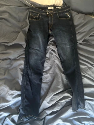 Mörkblå Replay Anbass slim fit jeans - Säljer ett par mörkblå jeans från Replay, modell Anbass med slim fit passform. Jeansen har klassisk femficksdesign, kontrastsömmar och är tillverkade i ett stretchigt denimtyg för extra komfort. Perfekta för dig som gillar en smalare siluett.