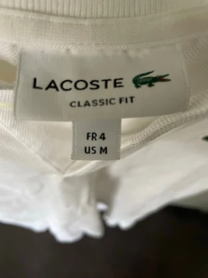 Vit pikétröja från Lacoste - Klassisk vit pikétröja från Lacoste med korta ärmar och krage. Tröjan har knappar vid halsen och den ikoniska gröna krokodilloggan på bröstet. Perfekt basic-plagg i garderoben för en clean och sportig look. Inga fläckar och passar dig väldigt fint