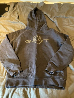 Svart hoodie från GANT med tryck - Svart hoodie från GANT med stort tryck på bröstet och huva. Klassisk modell med ribbade muddar och känguruficka. Perfekt för en avslappnad och stilren look. Tillverkad i mjukt bomullsmaterial.