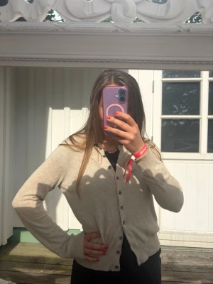 Beige kofta från Repeat easy knit - Snygg beige kofta från Repeat easy knit med rund halsringning och knäppning framtill. Koftan är tunnstickad och har långa ärmar samt ribbade muddar. Perfekt att slänga över en t-shirt eller linne för en chill look.