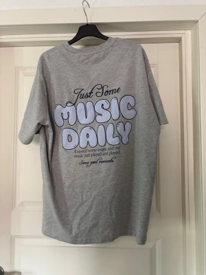 Grå t-shirt med tryck 'Music Daily' - Säljer en grå t-shirt med stort retroinspirerat tryck på ryggen där det står 'Just Some Music Daily' i ljuslila bokstäver. T-shirten har rund hals och korta ärmar. Perfekt för dig som gillar musik och vill ha en chill stil.