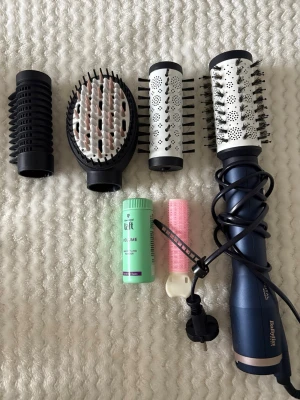 Babyliss varmluftsborste med tillbehör - Multifunktionell varmluftsborste från Babyliss i mörkblått med fyra utbytbara borsthuvuden, volympuder (använd endast en gång) och en rosa hårspole. Medföljer även en marin förvaringspåse. Perfekt för styling och volym till håret.