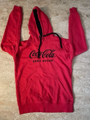 Röd Coca-Cola hoodie - Snygg röd hoodie med svart Coca-Cola Zero Sugar-tryck på bröstet. Klassisk känguruficka framtill, svart dragsko i huvan och mjukt material. Perfekt för dig som gillar streetwear och vill sticka ut med en ikonisk logga.