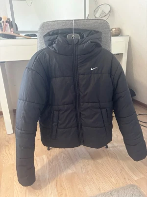 Svart pufferjacka från Nike - Snygg svart pufferjacka från Nike med huva och dragkedja framtill. Jackan har två fickor på sidorna och den klassiska vita Nike-loggan på bröstet. Perfekt för kalla vinterdagar och ger en sportig vibe.