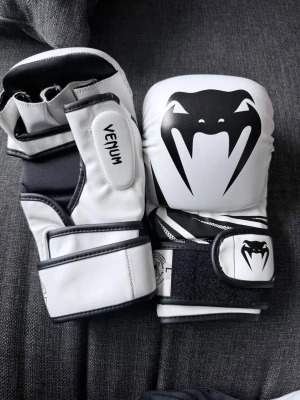 Venum vita MMA-handskar - Säljer ett par vita MMA-handskar från Venum med svart logga och detaljer. Handskarna har kardborreband för enkel justering och bra passform. Perfekta för träning inom kampsport eller sparring. Snygg och sportig design med tydliga svarta kontraster.