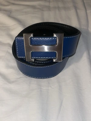 Blått skärp med stor H-spänne - Snyggt blått bälte i skinn med ett stort, silverfärgat H-format spänne. Bältet har vita sömmar längs kanterna som ger en clean kontrast. Perfekt accessoar för att lyfta din outfit.
