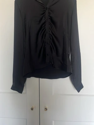 Svart blus med rynk framtill - Snygg svart blus i ett glansigt material med knappar och rynkdetaljer längs knäppningen framtill. Blusen har lång ärm och en något lös passform, perfekt till både jeans och kjol. Klassisk krage och mjukt tyg som ger en elegant look.