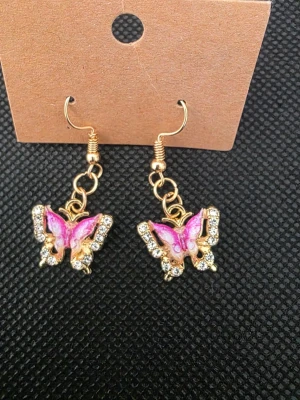 Guldiga örhängen med rosa fjärilar - Säljer ett par snygga örhängen i guldton med hängande fjärilar i rosa och vitt. Fjärilarna är dekorerade med glittrande stenar som ger extra bling. Perfekta för dig som gillar detaljer och färg. Krokarna är i metall och örhängena har en lekfull vibe.