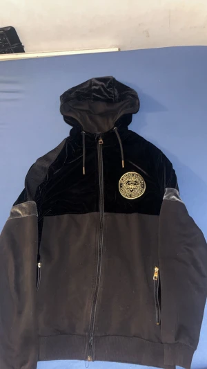 Svart hoodiejacka från Glorious Gangsta - Svart hoodiejacka med dragkedja och guldiga detaljer från Glorious Gangsta. Jackan har en stor huva med snörning, mjuk sammet på övre delen och ärmarna, samt ett stort guldigt emblem på bröstet. Fickor med guldfärgade dragkedjor och ribbad nederkant.