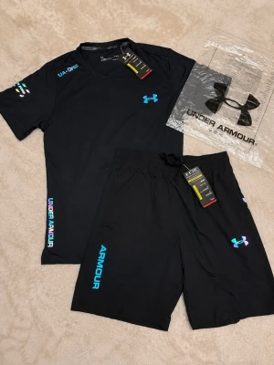 Svart träningsset från Under Armour - Snyggt svart träningsset från Under Armour med t-shirt och shorts. Setet har turkosa och regnbågsfärgade loggor och textdetaljer. Tillverkat i ett lätt och snabbtorkande funktionsmaterial som passar perfekt till träning och sport.