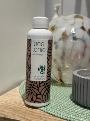 Face tonic med tea tree oil - Ungefär så mycket kvar som är vid mitt finger på bild 2. Fräsch ansiktstonic från Australian Bodycare med tea tree oil. Kommer i en vit plastflaska med brunt bladigt mönster och grön text. Perfekt för att rengöra och fräscha upp huden efter tvätt.