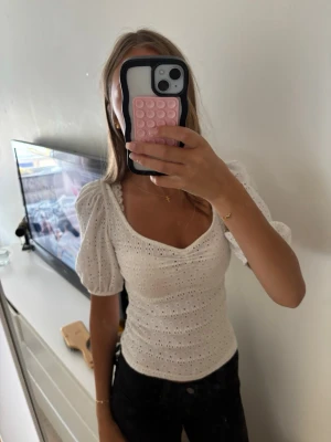 Vit broderad blus med puffärm - Söt vit blus med broderade detaljer och puffiga kortärmar. Toppen har v-ringning och sitter figurnära med fina spetsmönster över hela tyget. Perfekt för dig som vill ha en romantisk och trendig look.