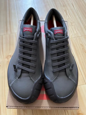 Gråa Camper sneakers i läder - Stilrena grå sneakers från Camper med lågt skaft och klassisk snörning. Skorna är tillverkade i slätt läder med robust gummisula och har röda detaljer på plösen och innersulan. Perfekta för dig som gillar minimalistisk och modern design.