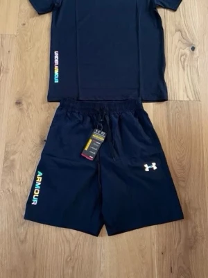 Svart träningsset från Under Armour - Snyggt svart träningsset från Under Armour med t-shirt och shorts. Båda plaggen har färgglada logotexter och klassisk UA-logga på shortsen. Perfekt för gymmet eller löprundan. Materialet är lätt och andas, vilket gör det bekvämt under träning.