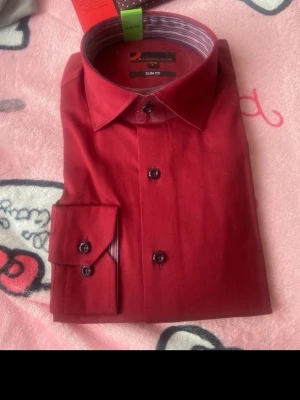 Röd slim fit skjorta från Dressmann - Snygg röd skjorta från Dressmann i slim fit-modell. Skjortan har klassisk krage, långa ärmar och knappar framtill. Perfekt för dig som vill ha en stilren och modern look. Tillverkad i mjukt bomullstyg för skön känsla.