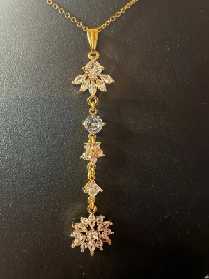 Guldigt halsband med gnistrande stenar - Elegant halsband i guldton med lång hängande berlock dekorerad med flera gnistrande, klara stenar i olika former. Berlocken har blommiga och geometriska detaljer som ger ett lyxigt och unikt intryck. Perfekt för dig som gillar att sticka ut med accessoarer.