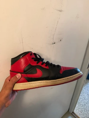 Jordan 1 mid bannad - Snygga sneakers i svart och rött läder med klassisk siluett och svart snörning. Skon har en vit sula med röd yttersula och ikonisk logga på sidan. Perfekt för dig som gillar streetwear och vill sticka ut med en retroinspirerad look.