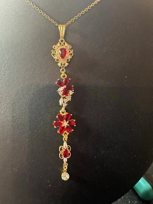 Guldhalsband med röda stenar och blommor - Unikt halsband i guldfärgad metall med hängande dekoration av röda och klara stenar i blomform. Kedjan är tunn och hänget har detaljerade utsmyckningar och gnistrande stenar som ger ett lyxigt intryck. Perfekt för dig som gillar statement-smycken och färgstarka accessoarer.
