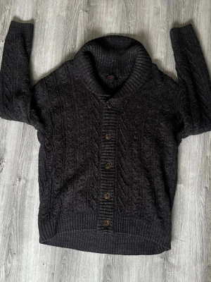 Indicode jeans stickad kofta  - Mysig mörkgrå kofta från indicode jeans med kabelstickat mönster och stora ribbade muddar. Koftan har en bred sjalkrage och knäpps med fem bruna knappar framtill. Den har inga tecken på användning. Hör av er vid frågor, allt gott! 