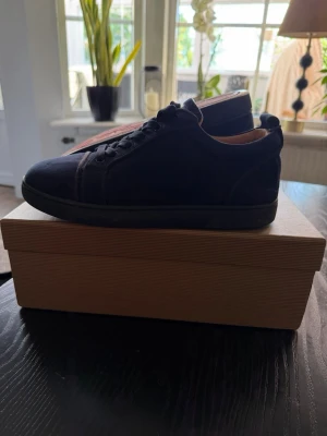 Mörkblå sneakers från Christian Louboutin - Snygga mörkblå sneakers från Christian Louboutin med klassisk låg profil och ikonisk röd sula. Skorna har ovandel i mocka och textil, matchande mörkblå snörning och diskreta sömdetaljer. 
