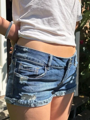 Lågmidjade blå jeansshorts - Säljer ett par lågmidjade blå jeansshorts med slitna detaljer. Shortsen har klassiska fickor fram och bak samt dragkedja och knapp. Super fint skick och perfekta nu i sommar! Knappt använda🥰 