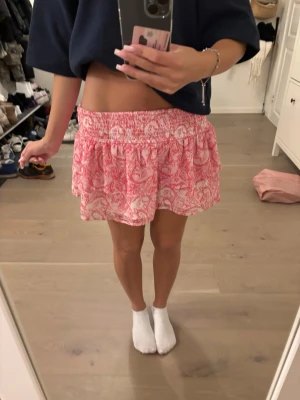 Rosa och vit kortkjol med volang - Supersöt kortkjol i rosa och vitt med volang och smock i midjan. Kjolen har ett blommigt mönster och är perfekt för varma dagar. Tillverkad i ett lätt och skönt material som känns luftigt. OBS HAR VIKT UPP DEN PÅ BILDEN. Storlek 152