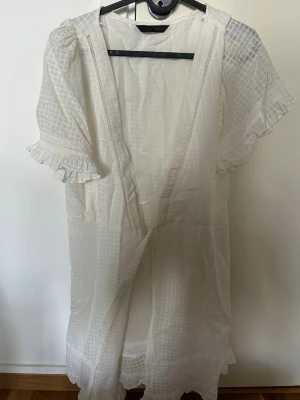 Vit klänning från Zara med volangärm - Luftig vit klänning som morgonrocks modell från Zara i lätt, transparent material med diskret rutmönster. Modellen har korta volangärmar och spetsdetaljer längs kanterna. Perfekt för chill dagar hemma eller som ett extra lager över pyjamasen.