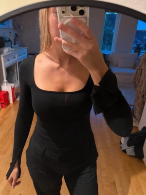 Svart långärmad topp med fyrkantig ringning - Snygg svart långärmad topp med fyrkantig ringning och figurnära passform. Ärmarna har en unik detalj med slits vid handlederna som ger en cool touch. Perfekt att styla med både jeans och kjol för en trendig look.