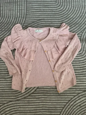 Ny Ljusrosa kofta med volangdetalj - Supersöt ljusrosa kofta med stora volanger över axlarna och dekorativa hålmönster på ärmar och framstycke. Koftan har rund halsringning, ribbade muddar och knäpps med små knappar framtill. Perfekt för dig som gillar en gullig och trendig stil.