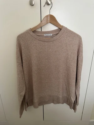 Beige stickad tröja Stradivarius - Mysig beige stickad tröja från Stradivarius med rund halsringning och ribbade muddar. Tröjan har en avslappnad passform och små slitsar i sidorna, perfekt för lager på lager. Enkel och stilren design som passar till det mesta.