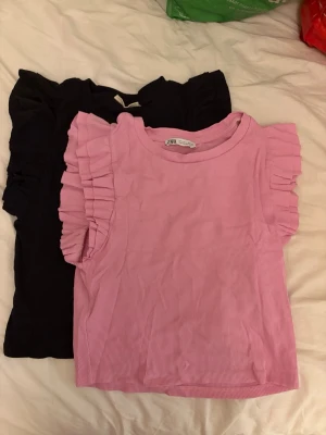 Rosa och svart volangtoppar från Zara - Två snygga toppar från Zara i storlek L, en i ljusrosa och en i svart. Båda har volangdetaljer längs ärmhålen och är ribbade. Perfekta för dig som vill ha en trendig och bekväm look. Svarta har fläck men det går säkert att tvätta bort 