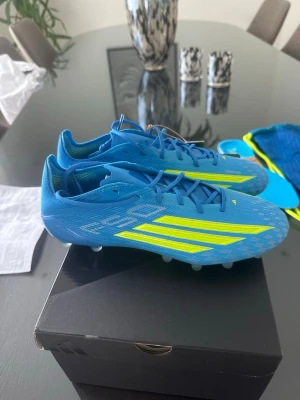 Blåa fotbollsskor med gula detaljer - Snygga blå fotbollsskor med skarpa gula ränder och mönster på sidorna. Skorna har snörning och låg profil, perfekt för snabba rörelser på planen. Tillverkade i syntetmaterial för lätt känsla och bra grepp på gräs.