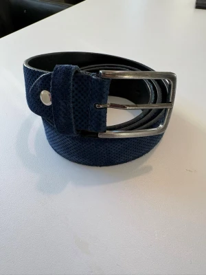 Mörkblått mockabälte i skinn - Snyggt mörkblått bälte i mocka med silverfärgat spänne. Bältet är tillverkat i Italien och har en stilren, klassisk design med diskret textur på ytan. Perfekt accessoar för att lyfta din outfit.