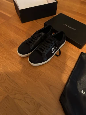 Svarta Saint Laurent sneakers - Snygga svarta sneakers från Saint Laurent med vit sula och handskriven logga på sidan. Skorna har klassisk låg profil, svarta skosnören och är tillverkade i en mix av läder och textil. Perfekta för dig som gillar stilrena och exklusiva detaljer.