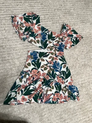 Blommig offshoulder kortklänning - Snygg kortklänning med offshoulder och volangärm. Vit botten med blommigt mönster i grönt, rosa och blått. Klänningen har en omlottliknande topp och markerad midja. Perfekt för sommarens alla tillfällen.