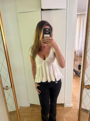 Vit spetsblus med volang och v-ringning - Söt vit blus med djup v-ringning och volangkant. Toppen är ärmlös och har fina spetsdetaljer som ger en romantisk känsla. Perfekt att styla med jeans eller kjol för en trendig look.
