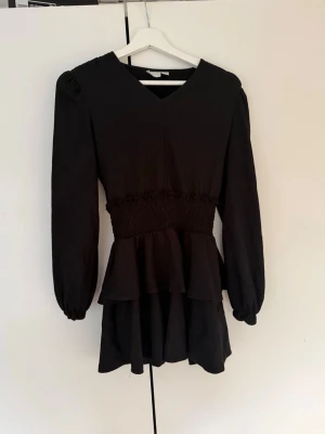 Svart kort klänning med volang - Snygg svart kort klänning med v-ringning, långa puffärmar och markerad midja. Klänningen har volangdetaljer nedtill och är perfekt för dig som gillar en feminin och trendig stil.