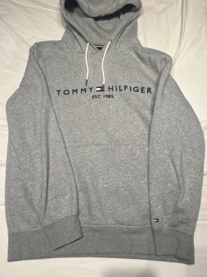 Grå hoodie från Tommy Hilfiger - Snygg grå hoodie från Tommy Hilfiger med klassisk logga och texten 'EST. 1985' broderat på bröstet. Tröjan har huva med vita dragsnören och ribbade muddar vid ärmar och nederkant. Tillverkad i mjukt bomullsmaterial för maximal komfort.