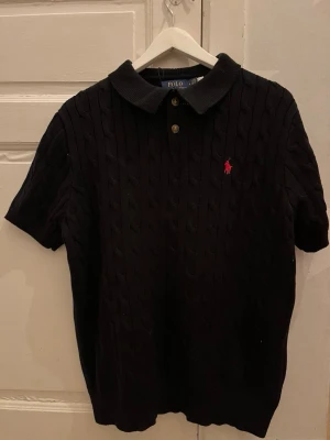 Svart kabelstickad pikétröja Polo Ralph Lauren - Kabelstickad svart pikétröja från Polo Ralph Lauren med korta ärmar, klassisk krage och två knappar framtill. Tröjan har den ikoniska röda logotypen broderad på bröstet och är tillverkad i mjuk bomull.Priset går att diskutera vid snabb affär, finn de frågor så är de bara o skriv 😊