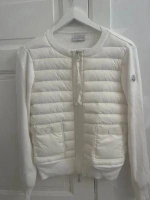 Vit quiltad cardigan från Moncler - Snygg vit cardiganjacka från Moncler med quiltad framsida och stickade ärmar. Jackan har två fickor framtill med tryckknappar, dragkedja och Moncler-logga på ärmen. Perfekt för lager-på-lager och en clean look. Priset går att diskutera vid snabb affär , finns de frågor att ställa så är de bara o skriv 😊