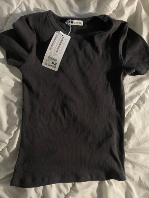 Svart ribbad t-shirt XS - En svart ribbad t-shirt i storlek XS med korta ärmar och rund hals. T-shirten har en enkel och stilren design som passar till det mesta. Materialet är mjukt och stretchigt för en bekväm passform.