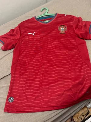 Röd fotbollströja Puma Portugal - Snygg röd fotbollströja från Puma med Portugals landslagsemblem på bröstet. Tröjan har blå detaljer vid krage och ärmslut samt diskreta vågmönster över hela tyget. Tillverkad i lätt och ventilerande material, perfekt för träning eller match.