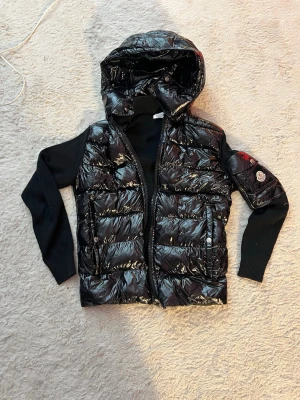 Svart pufferjacka med huva Moncler - Snygg svart pufferjacka från Moncler med glansig finish, huva och stickade svarta ärmar. Jackan har dragkedja framtill, två fickor med knappar och Moncler-logga på ärmen. Perfekt för kalla vinterdagar och ger en cool streetstil.