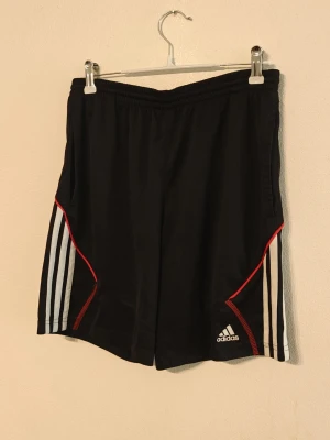 Adidas träningsshorts  - Säljer ett par svarta Adidas shorts med vita ränder längs sidorna och röda detaljer. Shortsen har elastisk midja och är tillverkade i lätt polyester, perfekta för träning eller sportiga dagar. Adidas-logga på vänster ben. Storlek M för barn.