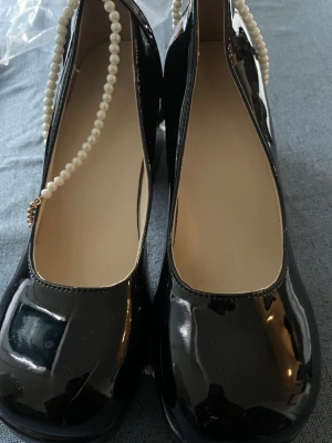 Svarta lackpumps med pärlband - Säljer ett par svarta pumps i glansigt lackmaterial med rund tå och chunky klack. Skorna har ett dekorativt pärlband runt vristen som ger en extra elegant touch. Perfekta för dig som vill sticka ut lite extra.