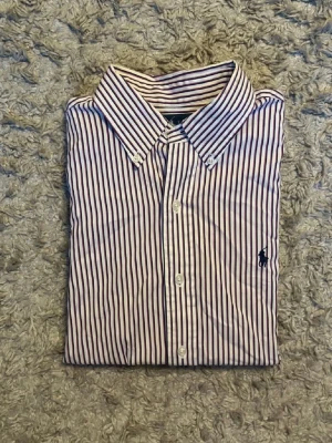 Ralph lauren skjorta - Klassisk ralph lauren med smala vertikala ränder i vitt och blått samt en broderad blå logga på bröstet. Skjortan har knappar framtill, button-down krage och långa ärmar. Perfekt för en clean och stilren look.