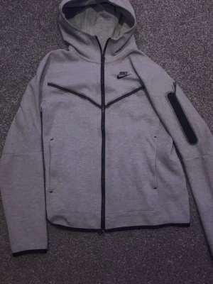 Grå Nike Tech Fleece hoodie - Grå Nike Tech Fleece hoodie med svarta detaljer. Mjuk, bekväm och stilren med hel dragkedja, huva och praktiska fickor. Perfekt för både vardag och träning.