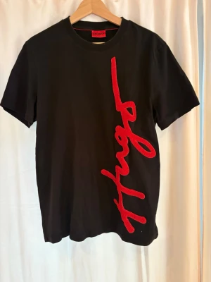 Svart t-shirt med röd Hugo-text - Snygg svart t-shirt från Hugo med stor röd text i sned design framtill. Klassisk rund halsringning och korta ärmar. Tillverkad i mjuk bomull som känns skön mot huden. Perfekt för en trendig och avslappnad stil.